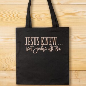 Puede incluir: Bolsa de tela negra con asas blancas y el texto "JESUS KNEW... but Judas ate too" en color marr&oacute;n claro.