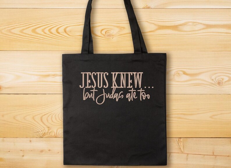 Puede incluir: Bolsa de tela negra con asas blancas y el texto "JESUS KNEW... but Judas ate too" en color marr&oacute;n claro.