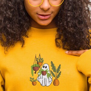 May include: A yellow sweatshirt with a white ghost holding a potted plant. The ghost is surrounded by other potted plants, including a hanging plant.