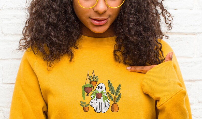 May include: A yellow sweatshirt with a white ghost holding a potted plant. The ghost is surrounded by other potted plants, including a hanging plant.