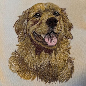 May include: Embroidered portrait of a Golden Retriever dog. The dog has brown fur, a black nose, and a pink tongue. The embroidery is detailed, capturing the dog's expression and fur texture. The background is a light cream colour.