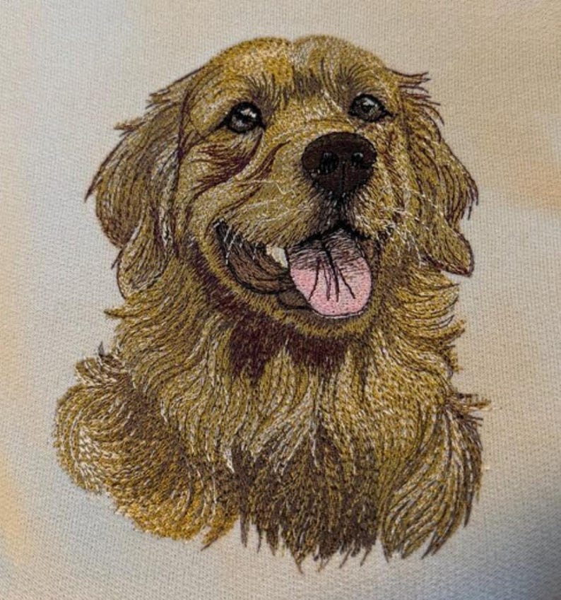 May include: Embroidered portrait of a Golden Retriever dog. The dog has brown fur, a black nose, and a pink tongue. The embroidery is detailed, capturing the dog's expression and fur texture. The background is a light cream colour.