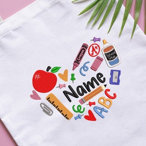 May include: White canvas tote bag with a heart-shaped design featuring school supplies like an apple, crayons, a pencil, a ruler, a glue stick, and the letters ABC. The text "Name" is in the center of the design.