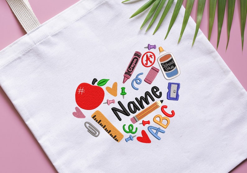 May include: White canvas tote bag with a heart-shaped design featuring school supplies like an apple, crayons, a pencil, a ruler, a glue stick, and the letters ABC. The text "Name" is in the center of the design.