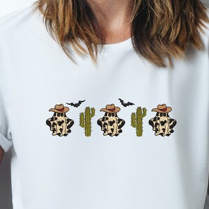 May include: White t-shirt with a graphic of three ghosts wearing cowboy hats and cacti. The ghosts are wearing a brown and white spotted pattern.
