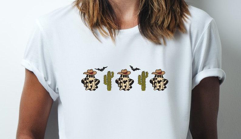 May include: White t-shirt with a graphic of three ghosts wearing cowboy hats and cacti. The ghosts are wearing a brown and white spotted pattern.