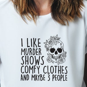 Op de afbeelding: Wit T-shirt met zwarte tekst die "Ik hou van moordseries, comfortabele kleding en misschien 3 mensen" luidt. Een zwart-witte schedel met bloemen is gecentreerd op het shirt.