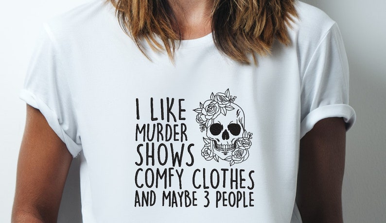 Op de afbeelding: Wit T-shirt met zwarte tekst die "Ik hou van moordseries, comfortabele kleding en misschien 3 mensen" luidt. Een zwart-witte schedel met bloemen is gecentreerd op het shirt.