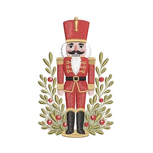 May include: A detailed embroidery of a red nutcracker soldier with a white beard and mustache, wearing a tall hat and black boots. The nutcracker is surrounded by a wreath of green leaves and red berries, perfect for holiday decor.