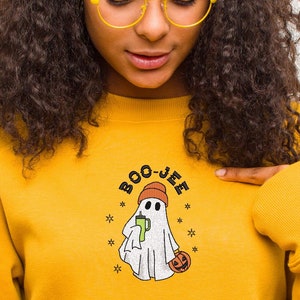May include: A yellow sweatshirt with a white ghost wearing a red hat holding a green drink and a pumpkin. The text "BOO-JEE" is above the ghost.