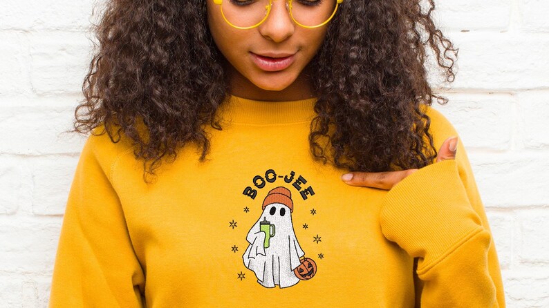 May include: A yellow sweatshirt with a white ghost wearing a red hat holding a green drink and a pumpkin. The text "BOO-JEE" is above the ghost.