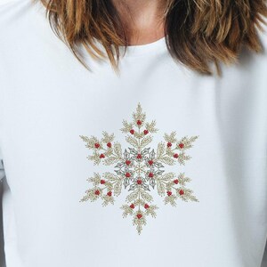 Puede incluir: Camiseta blanca con un dise&ntilde;o detallado de copo de nieve. El copo de nieve est&aacute; compuesto por hojas doradas y bayas rojas, con un centro plateado. El dise&ntilde;o est&aacute; centrado en el pecho de la camiseta.