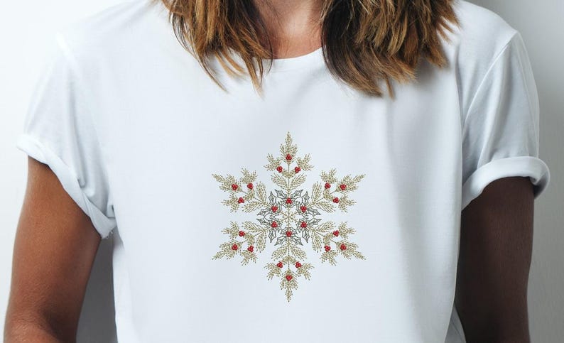 Puede incluir: Camiseta blanca con un dise&ntilde;o detallado de copo de nieve. El copo de nieve est&aacute; compuesto por hojas doradas y bayas rojas, con un centro plateado. El dise&ntilde;o est&aacute; centrado en el pecho de la camiseta.