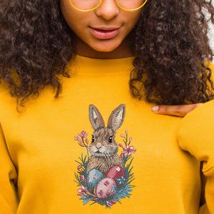 May include: A yellow sweatshirt with an embroidered design of a brown bunny rabbit holding Easter eggs surrounded by flowers.