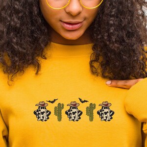 May include: A yellow sweatshirt with a design of four cow print ghosts wearing cowboy hats and cacti. The ghosts are wearing brown cowboy hats and have a black and white cow print pattern. The cacti are green and have brown stems. There are also two black bats flying above the ghosts.