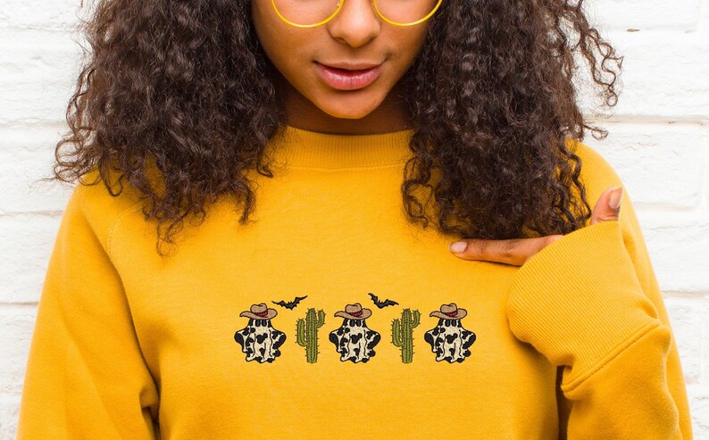 May include: A yellow sweatshirt with a design of four cow print ghosts wearing cowboy hats and cacti. The ghosts are wearing brown cowboy hats and have a black and white cow print pattern. The cacti are green and have brown stems. There are also two black bats flying above the ghosts.