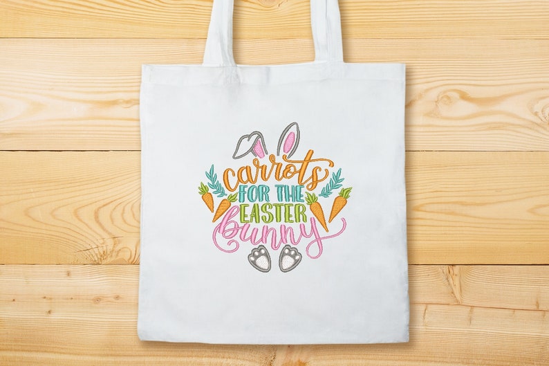 K&ouml;nnte beinhalten: Wei&szlig;e Einkaufstasche mit einem gestickten Design aus Karotten, Hasenohren und dem Text "Karotten f&uuml;r den Osterhasen".