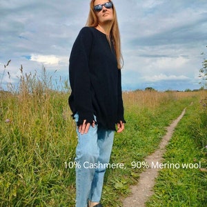 Könnte beinhalten: Eine Frau trägt einen schwarzen Pullover mit ausgefranstem Saum, blaue Jeans und schwarze Sandalen. Der Pullover besteht aus 10% Kaschmir und 90% Merinowolle. Die Frau steht auf einem Feld mit hohem Gras und einem Feldweg.