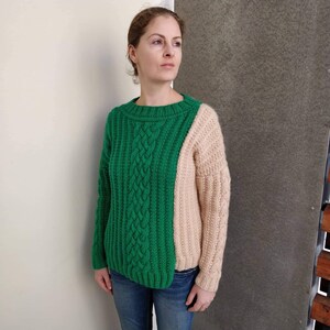 May include: A green and beige knitted jumper with a cable knit pattern on the green side. The jumper has a relaxed fit and a round neckline.