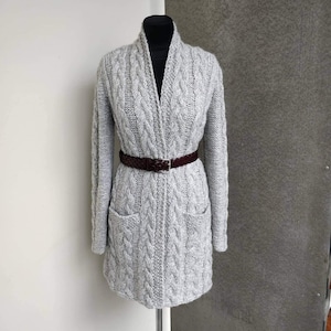May include: A light gray cable-knit cardigan with long sleeves and two front pockets. The cardigan is cinched at the waist with a brown leather belt. The garment is displayed on a black mannequin.