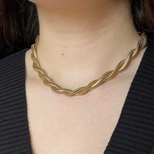 May include: A gold-toned chain necklace with a twisted design. The necklace is worn by a person with dark hair and a black shirt.