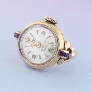 May include: A vintage gold-toned ring watch with a white face and black numerals. The watch has a small crown and is accented with two purple gemstones. The watch face reads "ELGA" and "INCABLOC".
