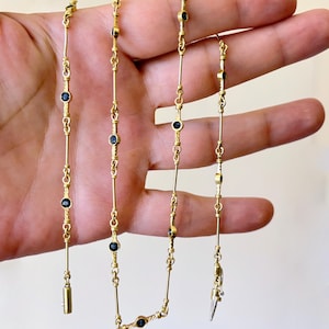 May include: A gold chain necklace featuring a series of evenly spaced, small, round, dark blue gemstones set in gold bezels. The chain consists of alternating straight gold bars and textured, twisted gold links connecting each gemstone setting.