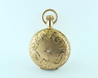 May include: A gold pocket watch with intricate floral and scrollwork engravings. A small diamond is set in the center of the watch face. The watch has a round shape with a loop at the top for a chain.