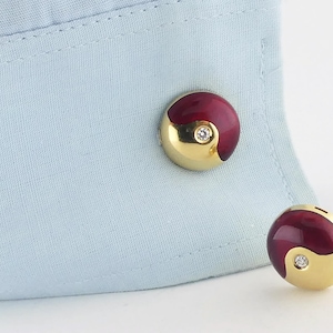 May include: A pair of gold-tone cufflinks with a yin-yang design. The cufflinks feature a deep red enamel and a gold-tone finish, with a small diamond accent in the center of each. The cufflinks are displayed on a light blue shirt cuff.