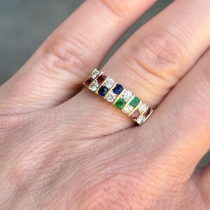 May include: A gold ring with a row of alternating colored gemstones. The gemstones include red, blue, white, and green stones. The ring is set with multiple small stones, creating a colorful and eye-catching design.