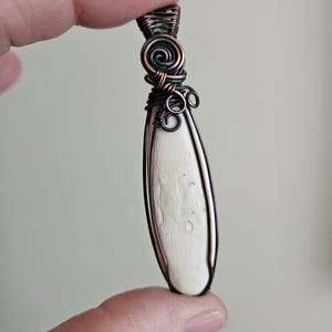 May include: A long, oval-shaped pendant featuring a smooth, off-white stone center encased in dark, oxidized copper wire. The wire is intricately wrapped in decorative spirals and coils at the top, forming a secure bail for hanging the piece.