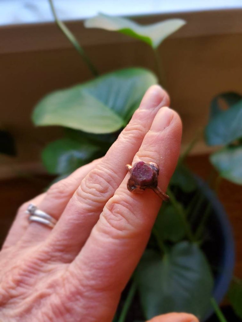 Op de afbeelding: Een koperkleurige ring met een grote, ruw geslepen, donkerrode edelsteen. De ring wordt aan een vinger gedragen, met een zilveren ring zichtbaar aan een andere vinger. Groene bladeren op de achtergrond.