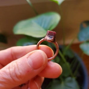 Op de afbeelding: Een handgemaakte koperen ring met een rechthoekige, roodbruine steen. De band van de ring heeft een eenvoudig, getextureerd ontwerp. De ring wordt vastgehouden tegen een achtergrond van groene bladeren en een vage achtergrond.