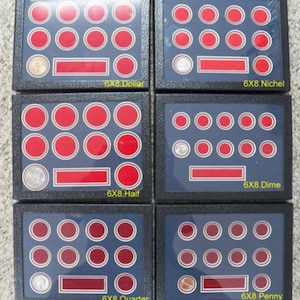 Op de afbeelding: Vier zwarte muntdisplays met rode fluwelen muntbewaarders. Elk display heeft een label met het munttype: Dollar, Nickel, Half, Dime, Quarter en Penny.