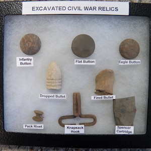 Może przedstawiać: Wytłoczona gablota z odnalezionymi relikwiami z wojny secesyjnej, w tym guzikami, kulami, hakiem do plecaka i nabojem Spencera. Gablota jest opisana jako "Excavated Civil War Relics".