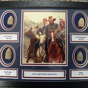 Könnte beinhalten: Eine gerahmte Präsentation von fünf Pistolenkugeln aus dem Amerikanischen Bürgerkrieg, jede in einem separaten runden Rahmen mit einem Etikett, das die Art der Kugel und das Kaliber identifiziert. Die Kugeln werden vor einem dunkelblauen Hintergrund mit einem Bild von konföderierten Soldaten zu Pferd in der Mitte ausgestellt.