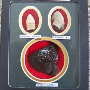 Op de afbeelding: Een zwarte vitrine met een wit label waarop "CIVIL WAR RELICS" staat. In de vitrine bevinden zich drie met rood fluweel beklede vakken, elk met een ander reliek uit de Amerikaanse Burgeroorlog. Het bovenste linker vak bevat een kogel die niet is afgevuurd. Het bovenste rechter vak bevat een kogel die is afgevuurd. Het onderste vak bevat een stuk van een granaatscherf.