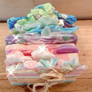 Puede incluir: Una pila de jabones artesanales coloridos, cada uno con un diseño único y una paleta de colores. Los jabones están atados con rafia y un lazo. Los colores incluyen rosa, azul, morado, verde y blanco.