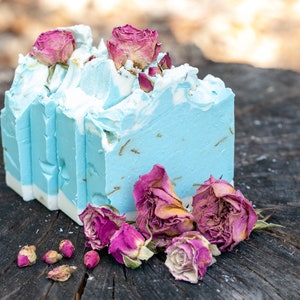 May include: A stack of handmade soap bars with a light blue and white swirl design, topped with dried pink roses. The soap is displayed on a weathered wooden surface, with additional rose buds scattered around.