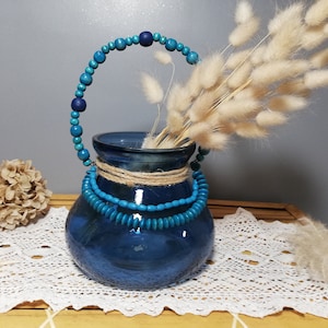 Peut inclure: Un vase en verre bleu avec une poignée en perles de bois et un accent de ficelle naturelle. Le vase est rempli d'herbe de pampa blanche séchée.