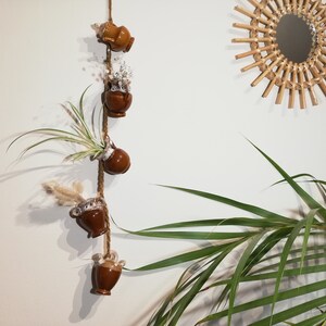 May include: A string of five brown ceramic pots hanging on a white wall. Each pot has a different plant or dried flower inside. A woven rattan sunburst mirror is on the wall to the right of the pots.