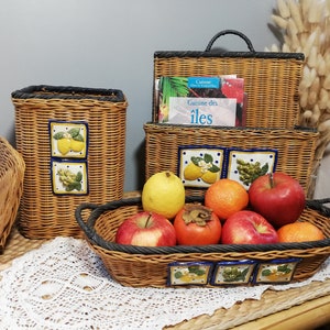 May include: A set of four wicker baskets with blue and white tile accents featuring lemons and grapes. The baskets are filled with fruit, including apples, oranges, persimmons, and lemons. The largest basket has a book titled "Cuisine des Iles" inside.