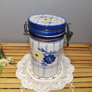 Puede incluir: Un tarro de cerámica blanco con un patrón a cuadros azul y blanco y un diseño floral. El tarro tiene una tapa de metal con un cierre.