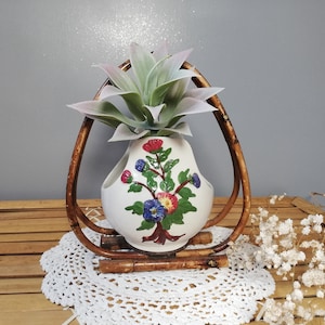 Peut inclure: Un vase vintage en céramique blanche avec un motif floral, encadré dans un cadre en bambou marron. Le vase contient une fausse plante succulente avec des feuilles vertes et roses. L'ensemble repose sur un napperon blanc et une surface en bois.