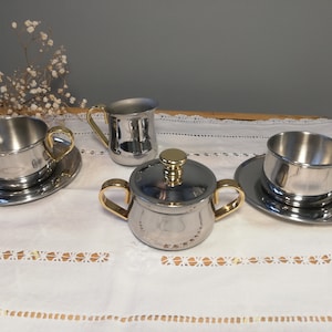May include: A set of four silver and gold coffee or tea service items. The set includes a creamer, a sugar bowl with a lid, and two cups and saucers.