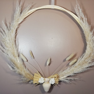 Peut inclure: Une couronne de pampas blanche avec un petit vase blanc et des fleurs artificielles au centre. La couronne est faite de matériaux naturels et est parfaite pour ajouter une touche d'élégance rustique à votre décoration intérieure.