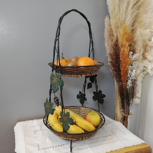 Peut inclure: Un panier à fruits à deux niveaux en fer forgé, avec une poignée. Le panier supérieur contient des oranges et des kiwis, le panier inférieur des bananes et une pomme. Les côtés sont ornés d'éléments décoratifs en forme de feuilles de vigne en métal.