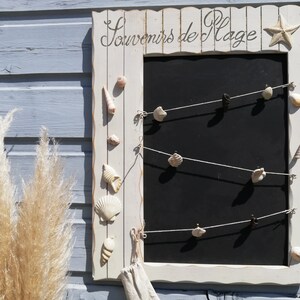 Peut inclure: Cadre en bois blanc avec un tableau noir à l'intérieur. Le cadre porte l'inscription "Souvenirs de Plage" et est décoré de coquillages.