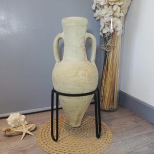 Pode incluir: Uma ânfora alta de cerâmica na cor creme com duas alças laterais, apoiada num suporte de arame de metal preto. O vaso apresenta uma base cónica e um pescoço largo, exibindo um acabamento de superfície texturizado e desgastado.