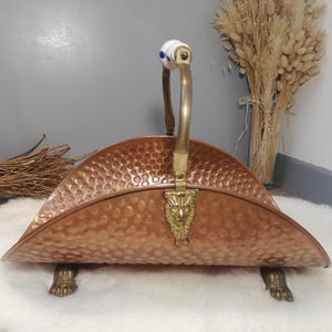 Peut inclure: Porte-revues cuivré avec une texture martelée. Il est doté d'une poignée en laiton avec un détail en céramique blanche et bleue et repose sur des pieds en forme de pattes de lion en laiton. Le porte-revues a un emblème de tête de lion décoratif.